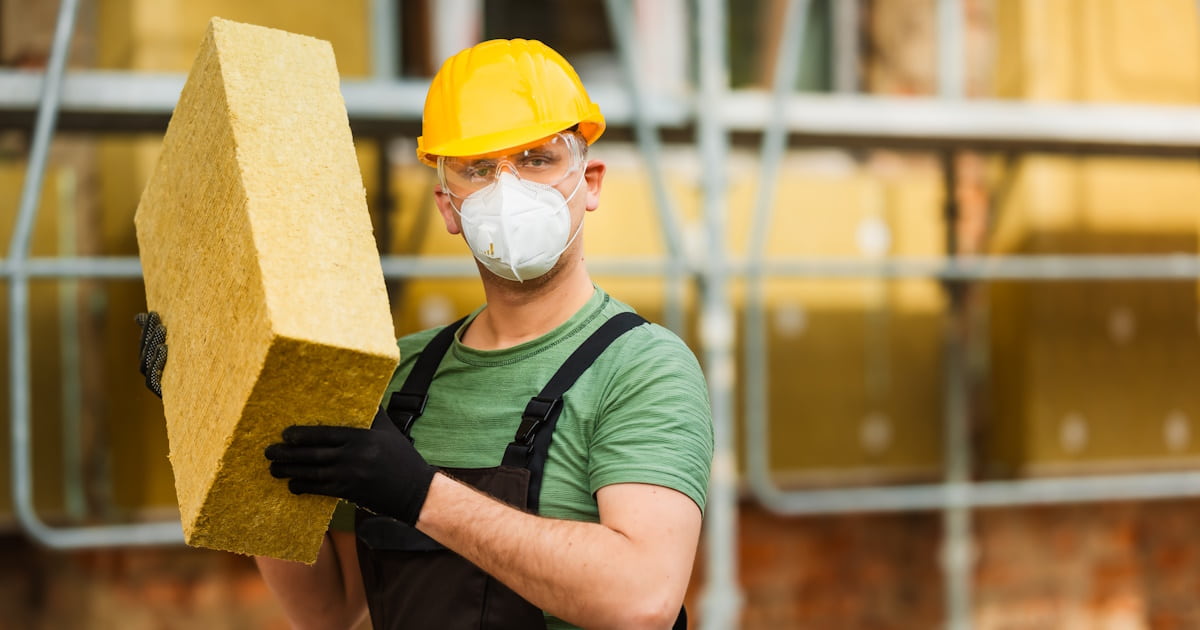 Een vakman draagt een blok isolatie waarmee een gevel geïsoleerd kan worden. Hij draagt een gele veiligheidshelm, handschoenen en een overall.