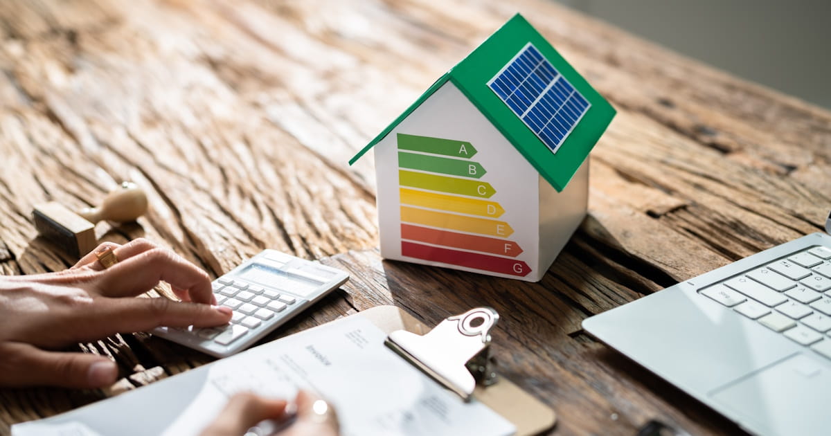 Een persoon berekent hoeveel de besparing van spouwmuurisolatie oplevert. Op tafel staat een huisje met energielabels, laptop, rekenmachine en klembord