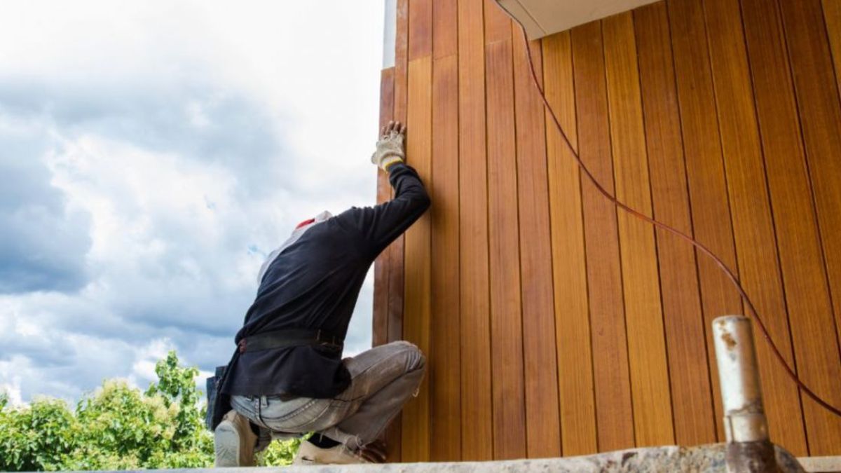Een vakman monteert thermowood gevelpanelen aan een muur, met een wolkenlucht op de achtergrond.