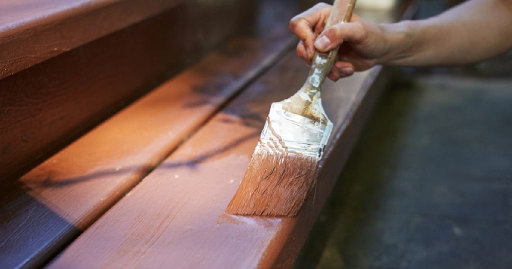 Een dameshand verft een houten trap met een kwast en donkere houtverf.
