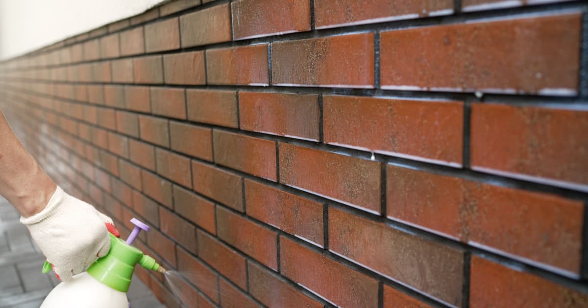 Een vakman brengt met een lagedrukspuit een waterafstotende impregneervloeistof aan op een bakstenen gevel, waarbij de natte behandelde zone duidelijk zichtbaar afsteekt tegen het droge metselwerk.