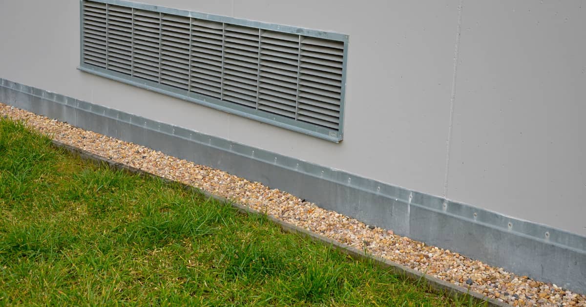 Rechthoekig metalen ventilatierooster met horizontale lamellen in een lichtgrijze buitenmuur vlak boven het maaiveld, met een grindstrook langs de gevel en groen gazon ervoor.