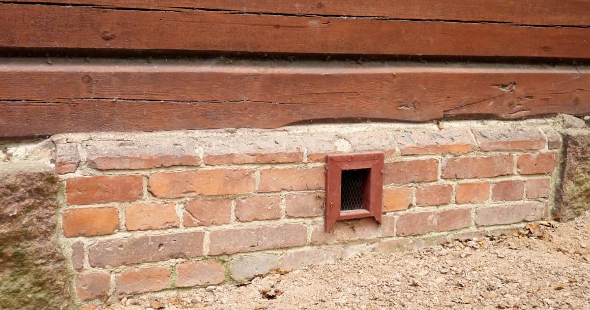 Een klein rood ventilatierooster met gaas zit in een lage bakstenen fundering onder een gevel van houten balken, met grind en droge bladeren op de grond ervoor.