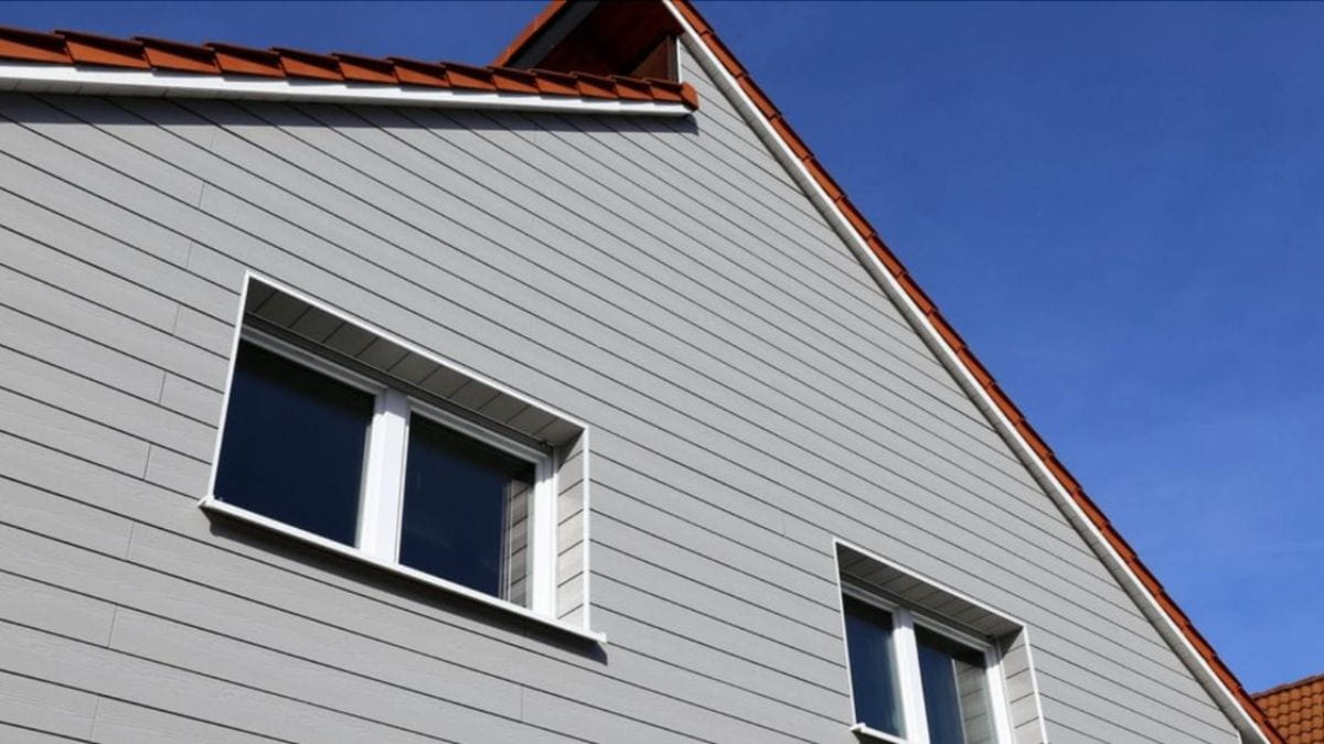 Woning met lichtgrijze vezelcement gevelbekleding (horizontale sidings), witte kozijnen en oranje dakpannen tegen een blauwe lucht.