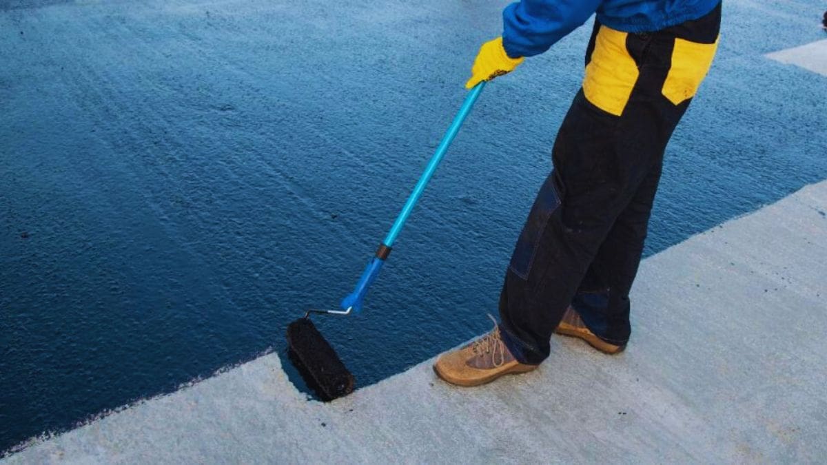 Een dakwerker brengt vloeibare bitumen dakbedekking aan op een betonnen dakvlak. Hij gebruikt een roller met telescoopsteel.
