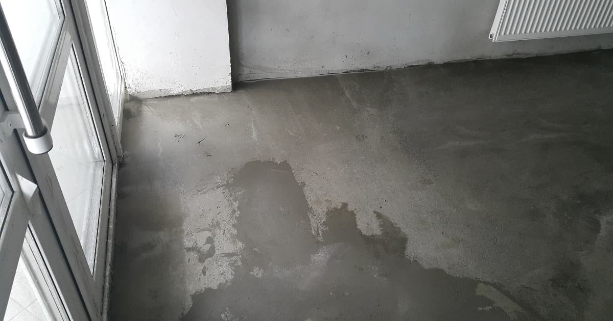 Een kale grijze betonvloer vertoont grote donkere natte plekken naast een openstaande glazen deur in een onafgewerkte ruimte met een radiator aan de muur.