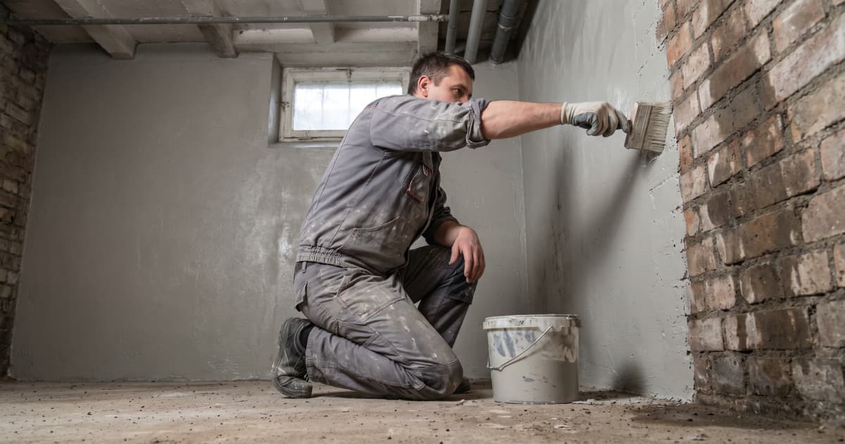 Een arbeider in grijze overall knielt in een kelder en brengt met een brede kwast een grijze waterdichte coating aan op een bakstenen muur, naast hem staat een emmer met coatingmateriaal.