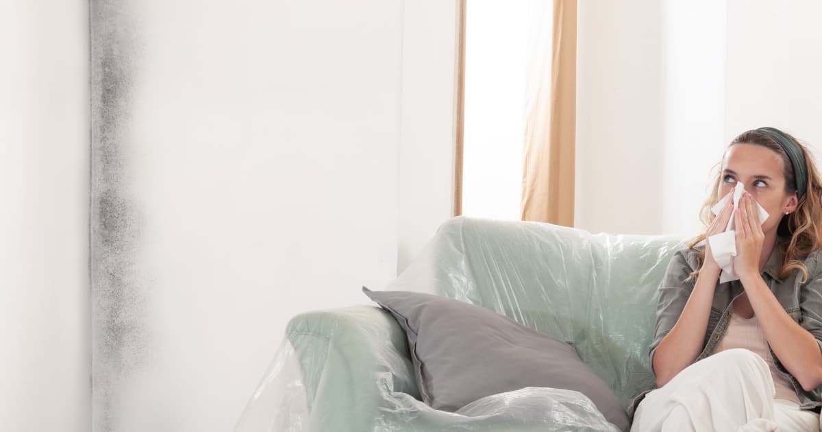 Een vrouw zit op een met plastic afgedekte bank en snuit haar neus in een zakdoek terwijl ze kijkt naar een witte muur met donkere schimmelvlekken in de hoek naast haar.