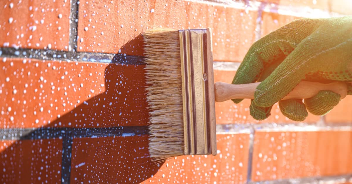 Een persoon met een groene handschoen brengt een middel aan op de bakstenen gevel