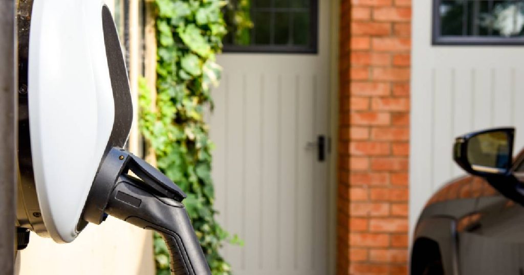 Een nieuwe residentiële woning waar, volgens de wetgeving in Nederland, een laadpaal of wallbox geïnstalleerd werd. Op de voorgrond de wallbox, op de achtergrond de gevel van de nieuwe woning en een elektrisch voertuig.