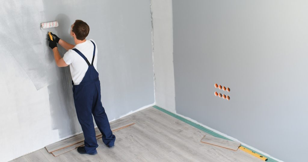 Een schilder verf een ruimte in een lichte kleur. De schilder draagt een overall en gebruikt een verfroller.