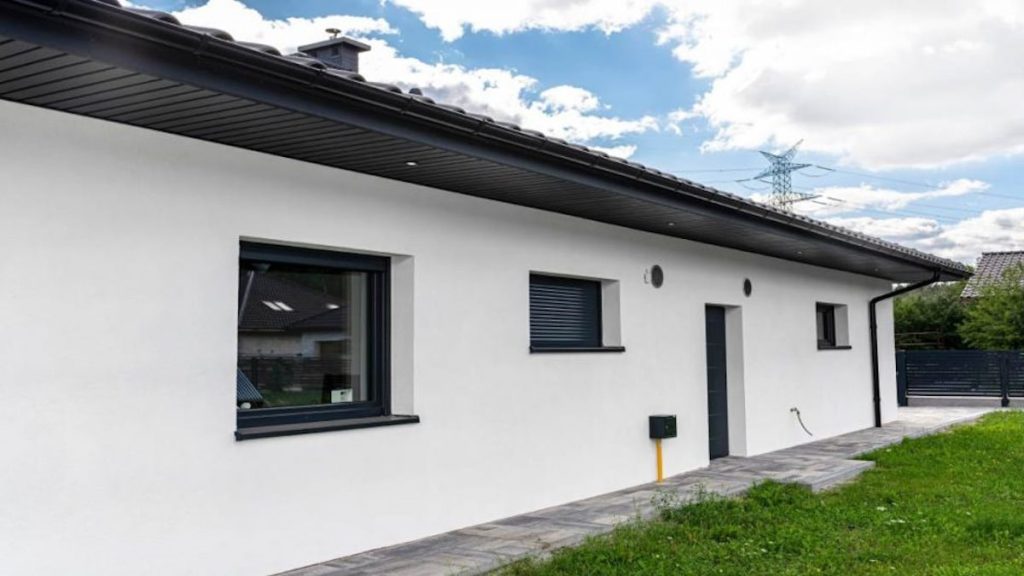 Een gelijkvloerse woning met een gevel van witte sierpleister, zwarte ramen en andere details. 
