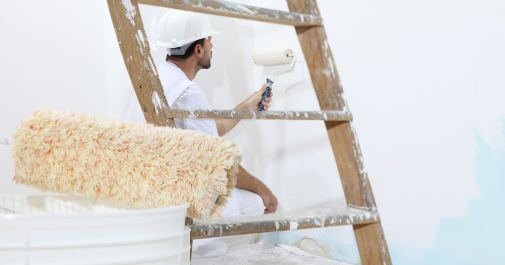Een schilder schildert een blauwe muur met witte verf. De schilder draagt een witte helm. Op de voorgrond zijn een verfroller en een ladder te zien.
