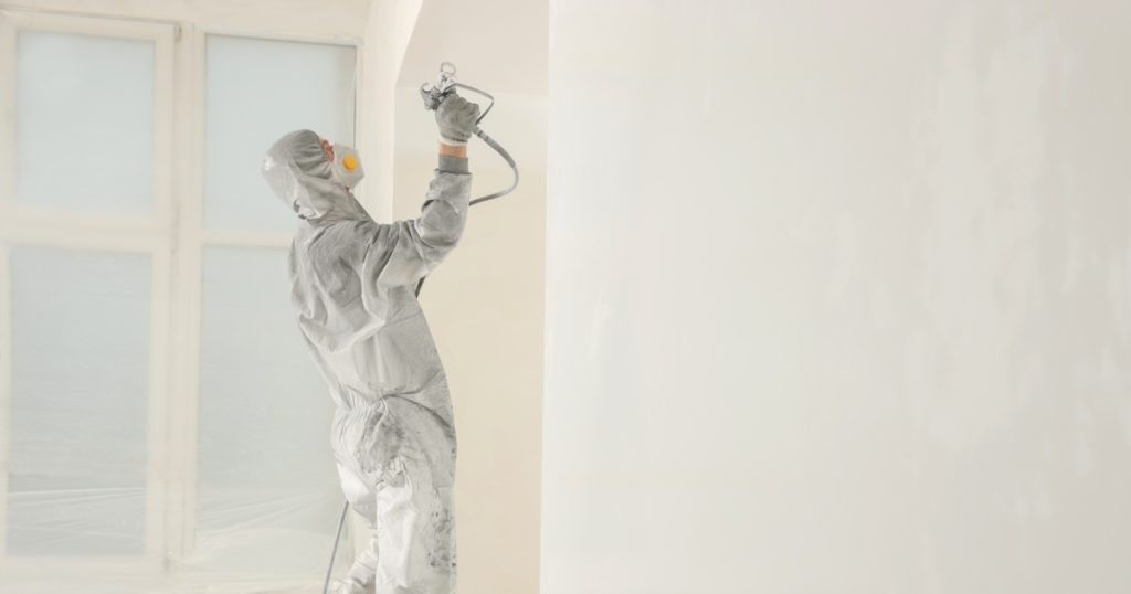 Een schilder spuit een muur in een lichte kleur. De schilder draagt beschermende kleding en een masker om te voorkomen dat hij de nevel inademt.