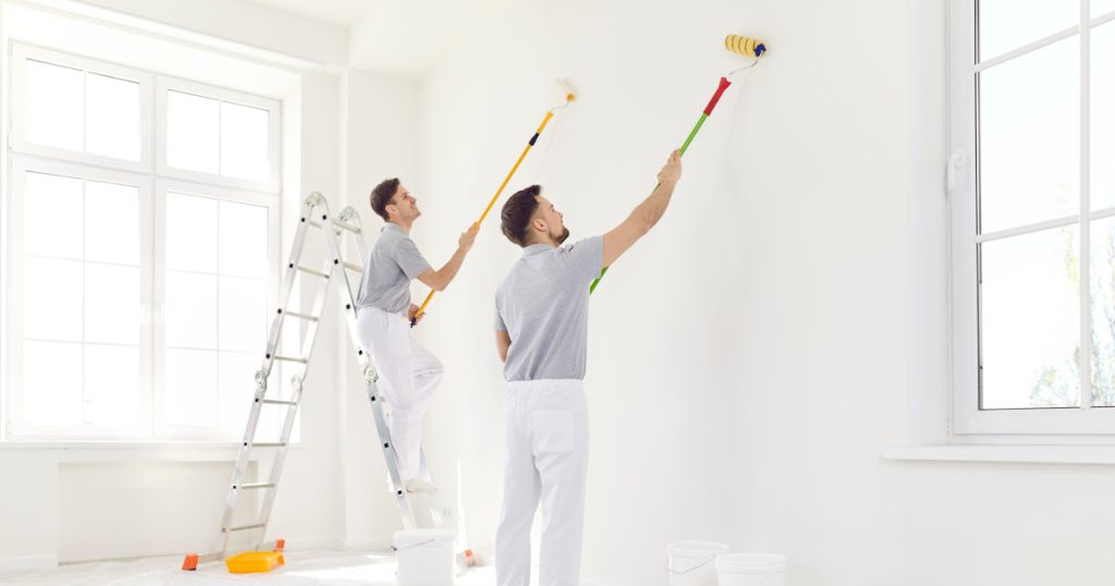 Twee schilders verven een wand met verfrollers. Een van de schilders staat op een ladder. De ruimte is licht.