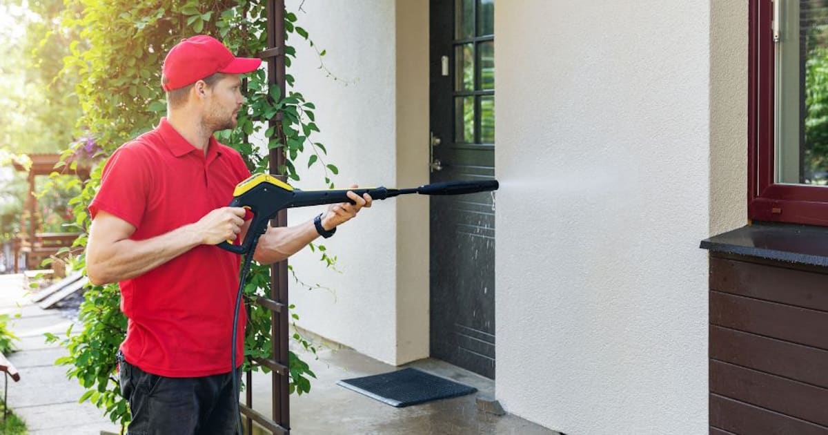 Een witte gevel wordt met een hogedrukreiniger gereinigd. De man op de foto draagt een rood shirt en een rode pet.